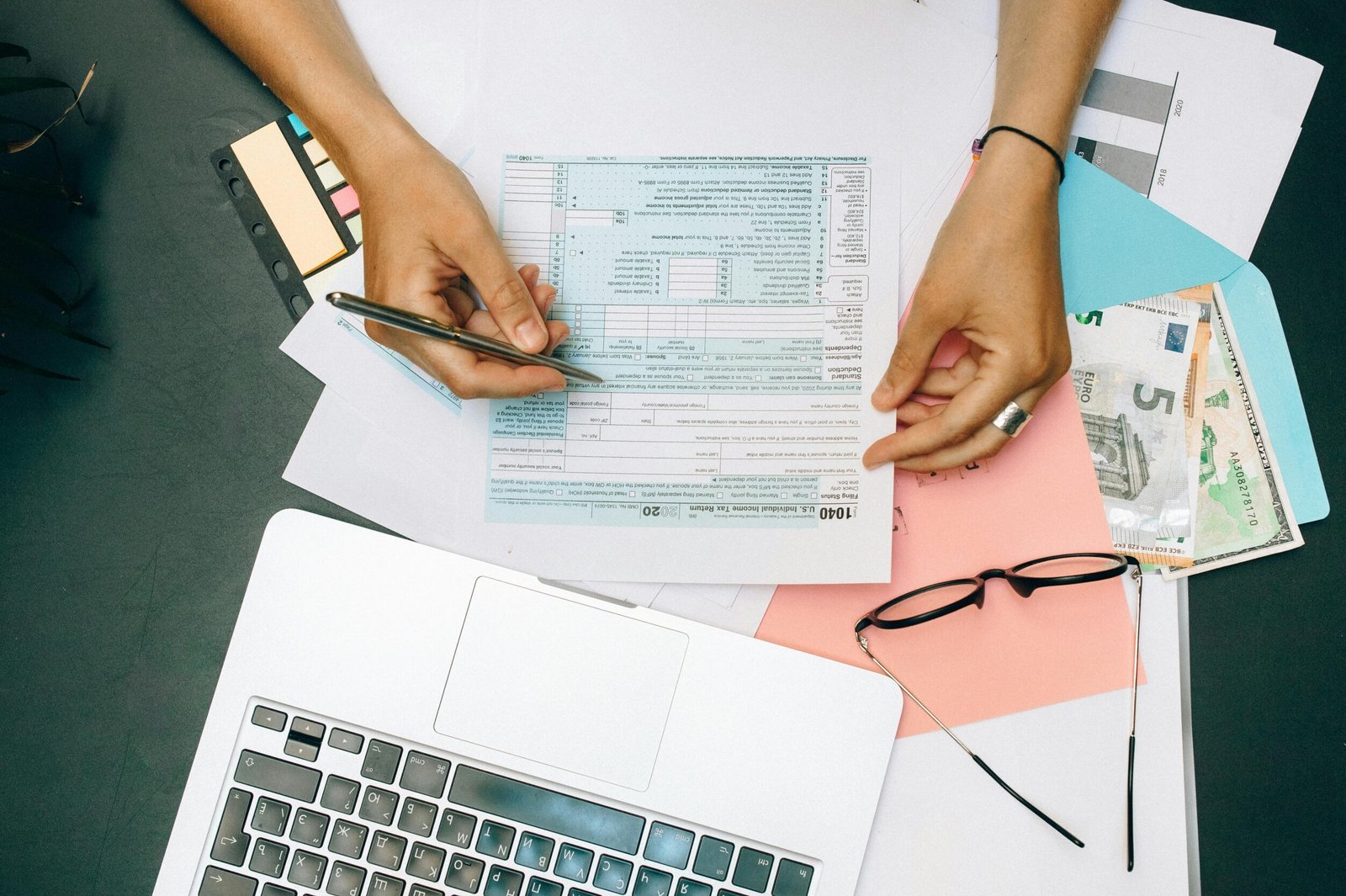 Hands writing on tax documents with laptop, glasses, and currency on desk.